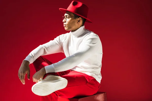 Vista lateral del elegante hombre de raza mixta en sombrero y gafas sentado y posando aislado en rojo - foto de stock