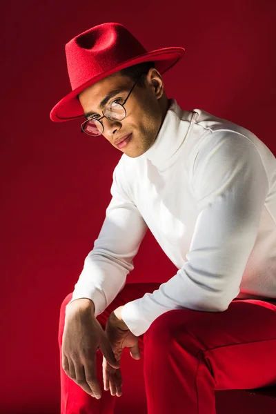 Stylish mixed race man in hat and glasses sitting, looking at camera and posing isolated on red — Stock Photo