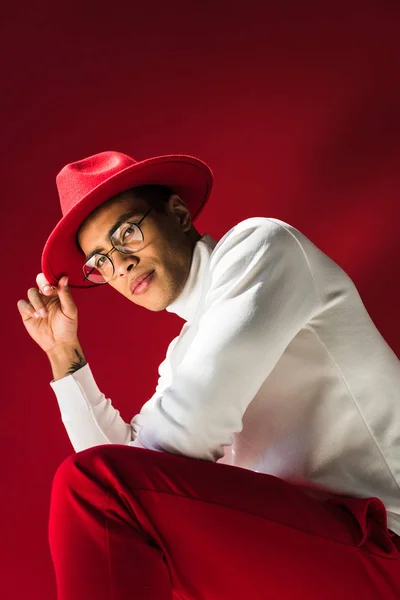 Vista de ángulo bajo de hombre elegante raza mixta en sombrero y gafas sentado y posando en rojo - foto de stock