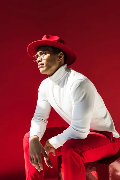 Uomo di razza mista alla moda in cappello e occhiali seduti e in posa isolata sul rosso — Foto stock