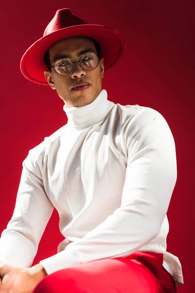 Fashionable mixed race man in hat and glasses sitting and posing isolated on red — Stock Photo