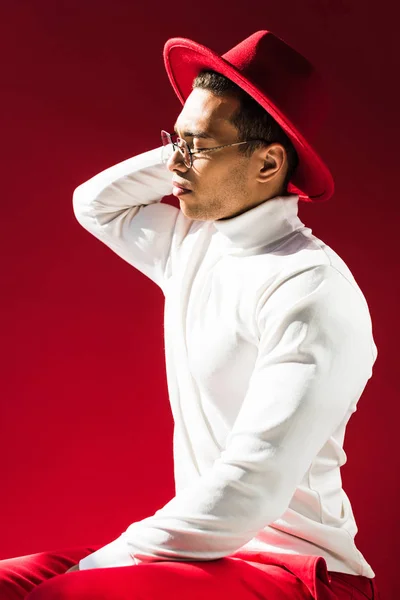 Elegante hombre de raza mixta en sombrero, cuello alto y gafas sentado y posando aislado en rojo - foto de stock