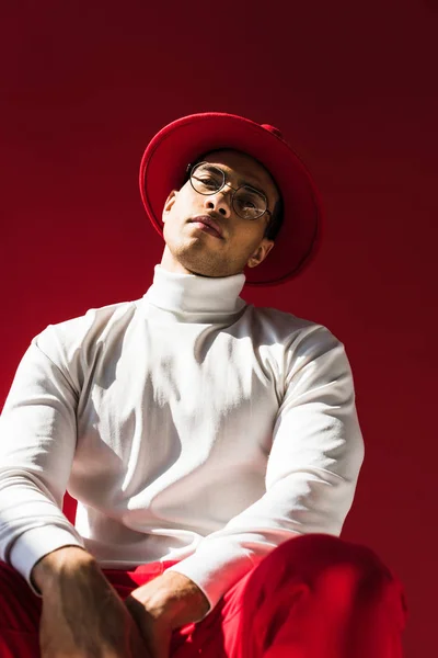 Stylish mixed race man in hat and glasses sitting, looking at camera and posing isolated on red — Stock Photo
