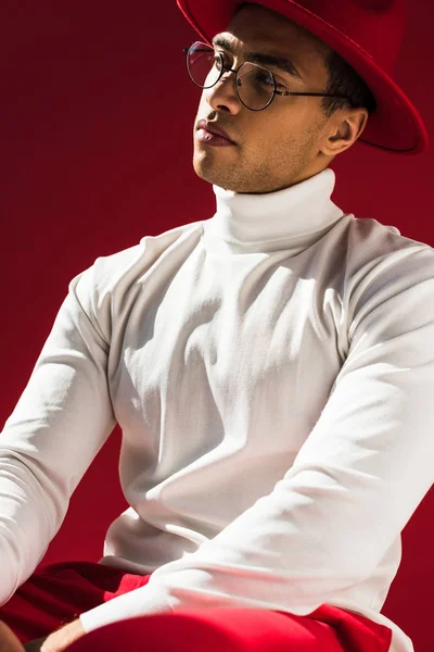 Confident stylish mixed race man in hat and glasses posing isolated on red — Stock Photo