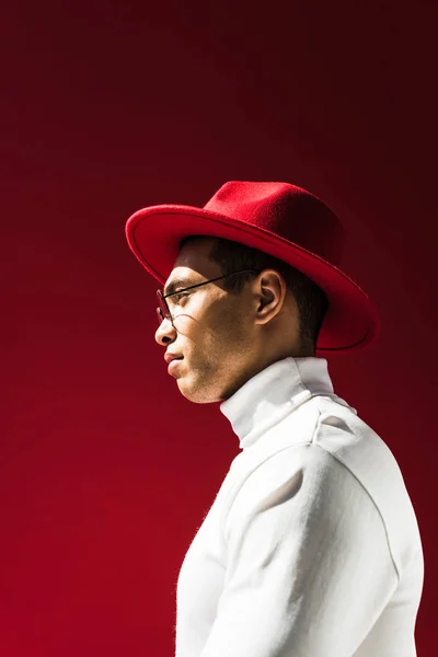 Side view of stylish mixed race man in hat and glasses posing isolated on red — Stock Photo