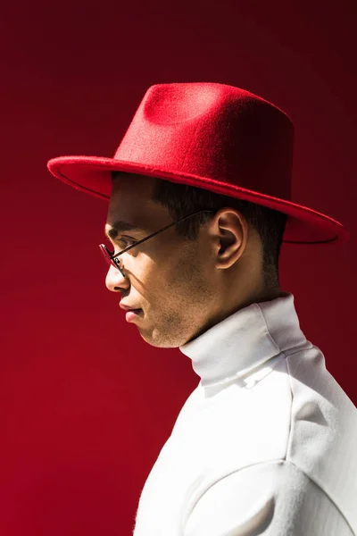 Vista laterale di elegante uomo razza mista in cappello e occhiali posa isolata sul rosso — Foto stock