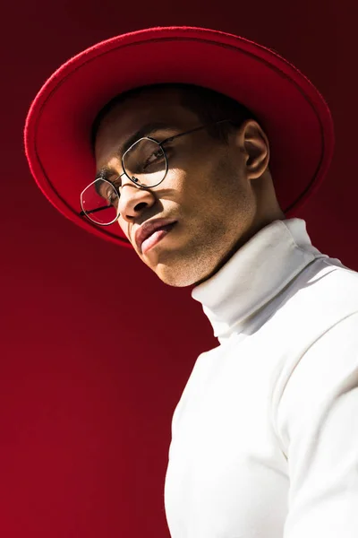 Stylish mixed race man in hat and glasses looking at camera while posing isolated on red — Stock Photo