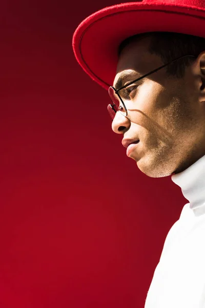 Вид збоку стильний змішаний гоночний чоловік в капелюсі і окулярах позує ізольовано на червоному з місцем для тексту — стокове фото