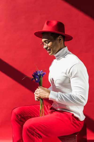 Homem de raça mista elegante em chapéu e óculos segurando flores e sorrindo no vermelho — Fotografia de Stock