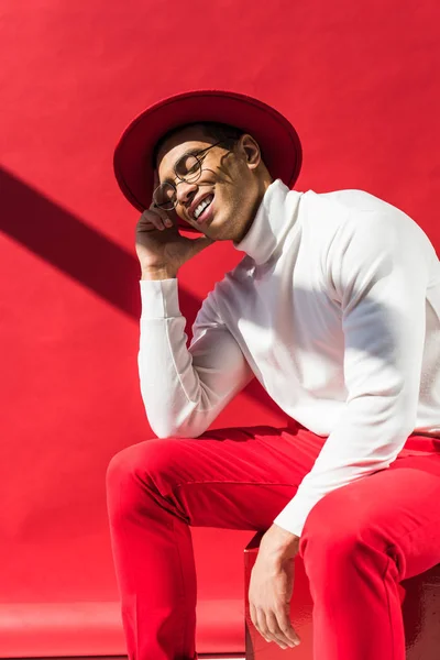 Stylish mixed race man in hat and glasses sitting, smiling and posing on red — Stock Photo