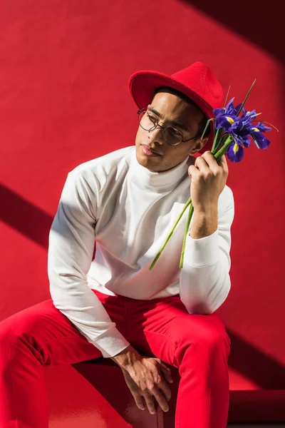 Stylish mixed race man in hat and glasses holding flowers on red — Stock Photo