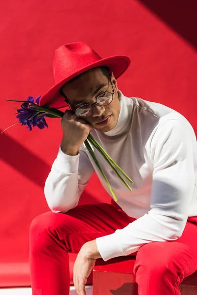 Stylish mixed race man in hat and glasses holding flowers on red — Stock Photo