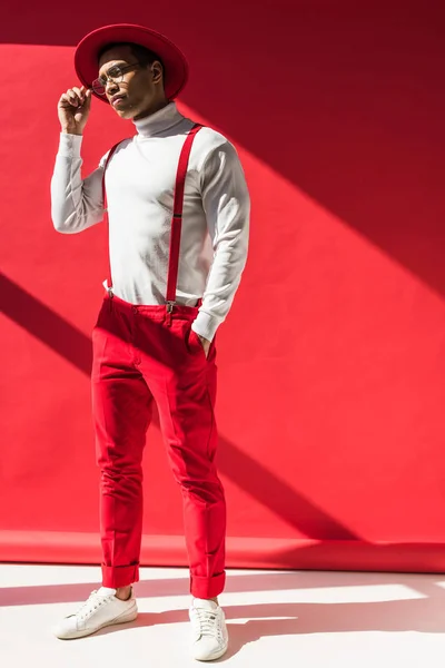 Модный человек смешанной расы в шляпе и подтяжки позируют на красном с копировальным пространством — стоковое фото