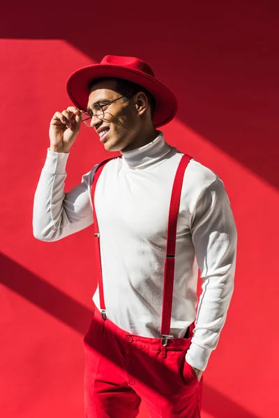 Hombre de raza mixta de moda en sombrero y tirantes sonriendo y posando en rojo - foto de stock