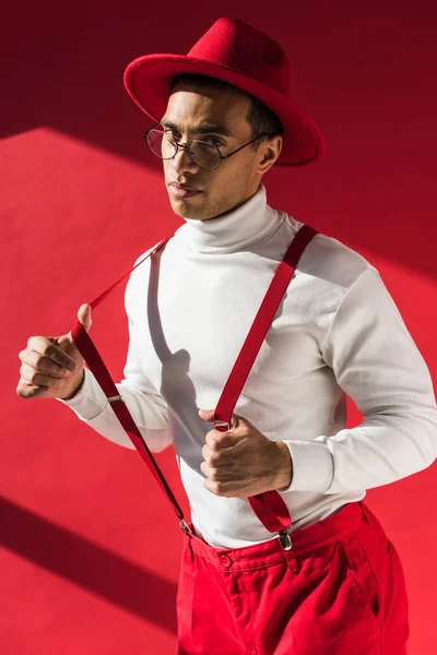 Fashionable mixed race man in hat and suspenders looking at camera while posing on red — Stock Photo