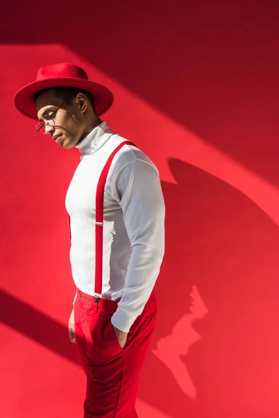 Fashionable mixed race man in hat, glasses and suspenders posing on red with copy space — Stock Photo