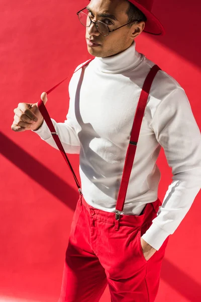 Fashionable mixed race man in hat and suspenders looking at camera while posing on red — Stock Photo