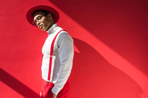 Fashionable mixed race man in hat and suspenders posing on red with copy space — Stock Photo