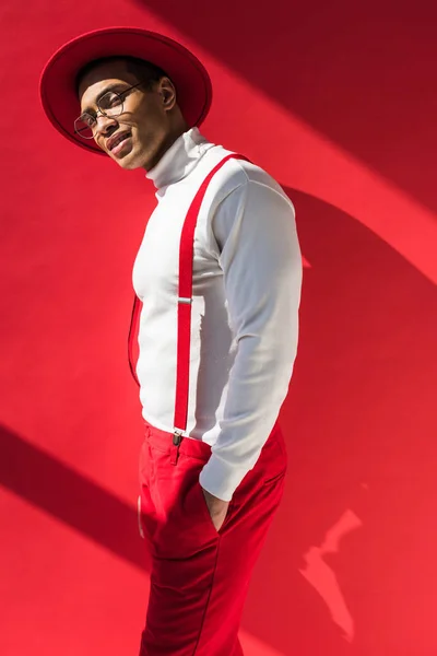 Stylish mixed race man in hat and suspenders looking at camera while posing on red — Stock Photo