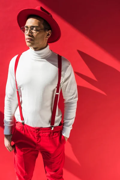 Stylish mixed race man in hat and suspenders posing with eyes closed on red — Stock Photo