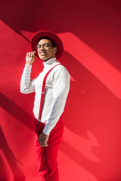 Homem de raça mista na moda em chapéu e suspensórios sorrindo e posando no vermelho com espaço de cópia — Fotografia de Stock