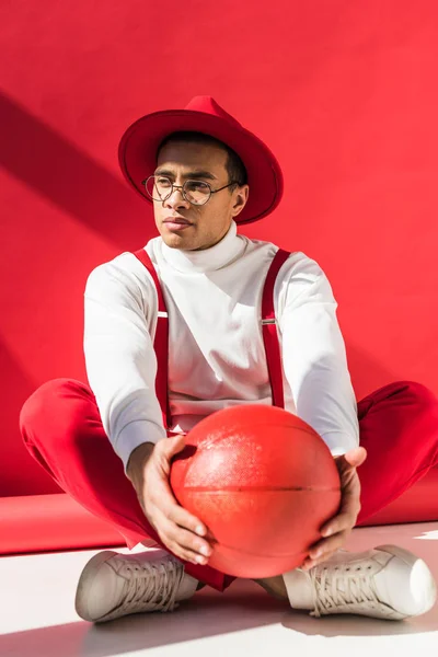 Elegante uomo di razza mista in cappello e bretelle seduto e in posa con pallacanestro sul rosso — Foto stock