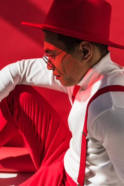 Side view of fashionable mixed race man in hat and suspenders posing on red — Stock Photo