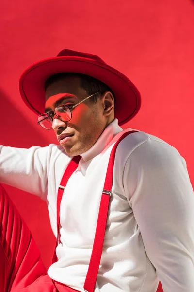 Hombre de raza mixta de moda en sombrero, gafas y tirantes posando en rojo - foto de stock