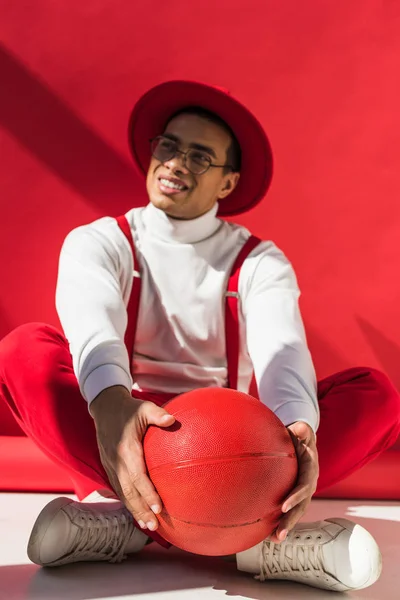 Stylish mixed race man in hat and suspenders sitting and posing with basketball on red — Stock Photo