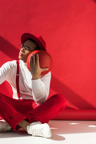 Elegante uomo di razza mista in cappello e bretelle seduto e in posa con pallacanestro sul rosso con spazio copia — Foto stock