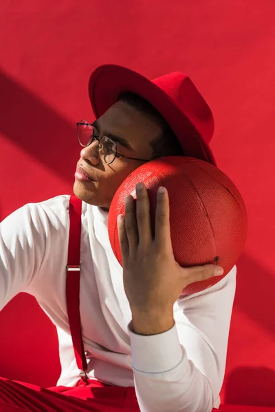 Stylish mixed race man in hat and suspenders posing with basketball on red — Stock Photo