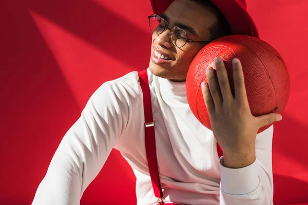 Elegante uomo di razza mista in cappello e bretelle sorridente mentre posa con pallacanestro sul rosso — Foto stock