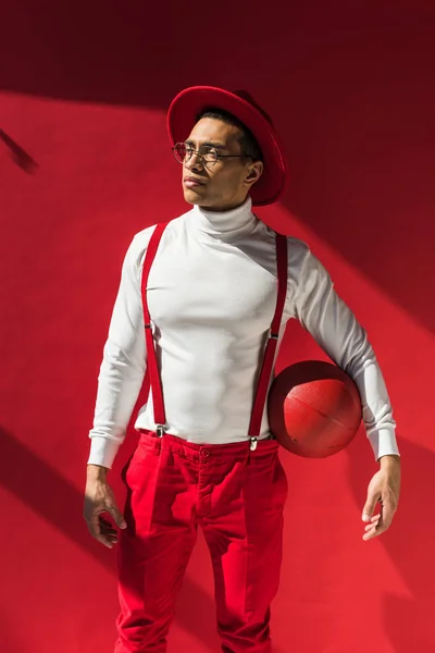 Stylish mixed race man in hat and suspenders posing with basketball on red — Stock Photo