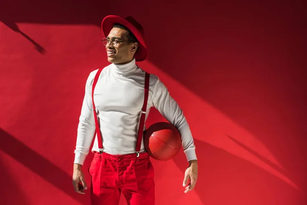 Stylish mixed race man in hat and suspenders posing with basketball on red with copy space — Stock Photo