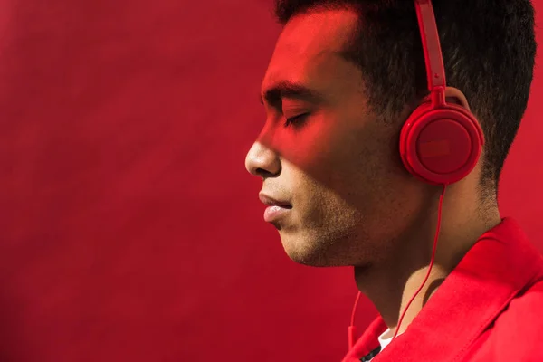 Side view of handsome mixed race man in headphones with eyes closed isolated on red with copy space — Stock Photo