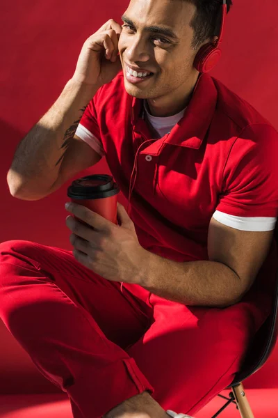 Stylish mixed race man in headphones sitting, holding coffee to go and talking on smartphone on red — Stock Photo