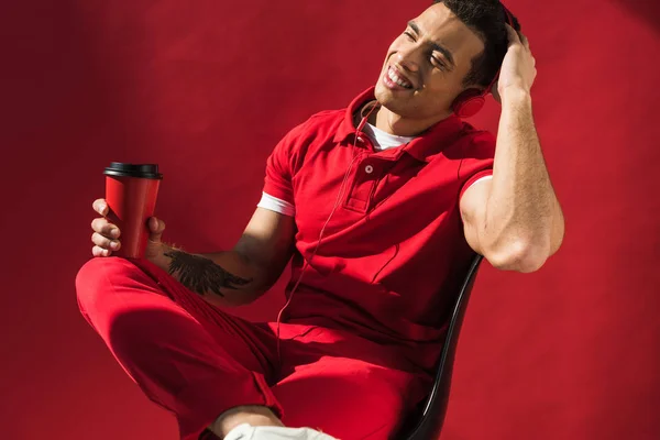 Stylish mixed race man in headphones sitting, holding coffee to go and smiling on red — Stock Photo