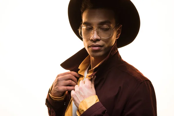 Stylish mixed race man in glasses and hat adjusting coat and posing isolated on white — Stock Photo