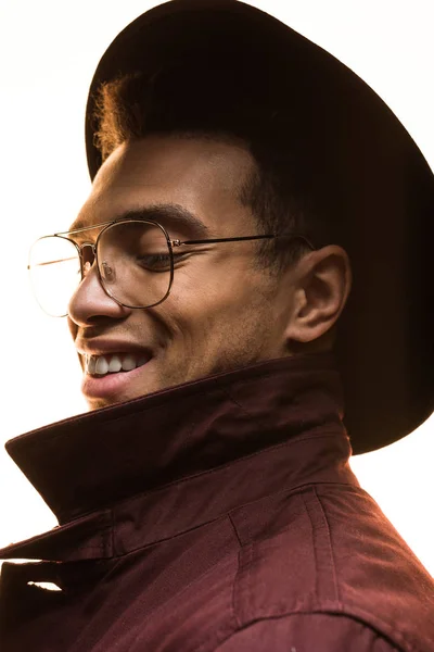 Elegante uomo misto razza in occhiali, cappello e cappotto sorridente e posa isolata su bianco — Foto stock