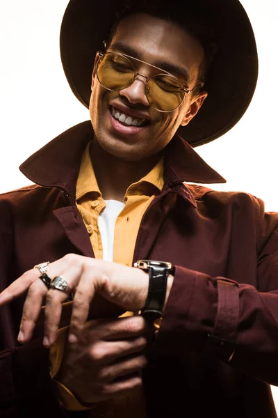 Stylish mixed race man in hat and sunglasses putting on watch and smiling isolated on white — Stock Photo