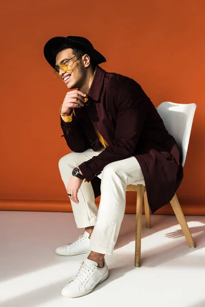 Elegante hombre de raza mixta en sombrero y gafas de sol sentado en la silla y sonriendo en naranja - foto de stock