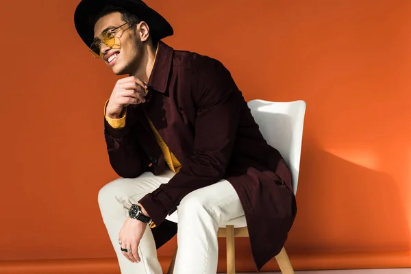 Elegante uomo di razza mista in cappello e occhiali da sole seduto sulla sedia e sorridente arancione — Foto stock