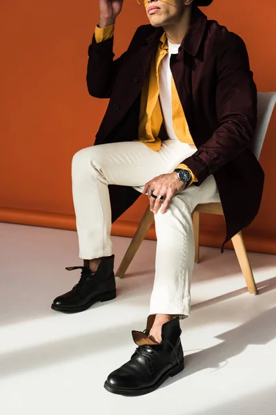 Cropped view of stylish mixed race man sitting and posing on orange — Stock Photo