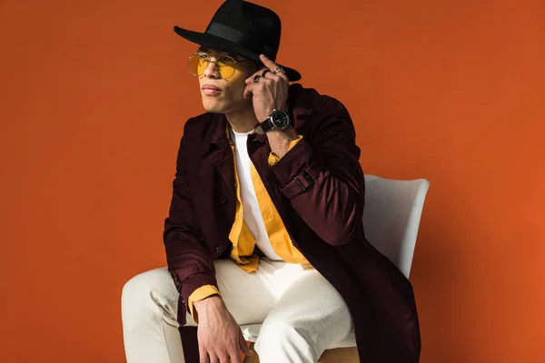 Elegante hombre de raza mixta en sombrero y gafas de sol sentado y posando aislado en naranja — Stock Photo