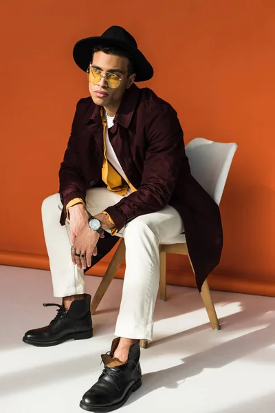 Uomo di razza mista alla moda in cappello e occhiali da sole seduto e in posa su arancione — Foto stock