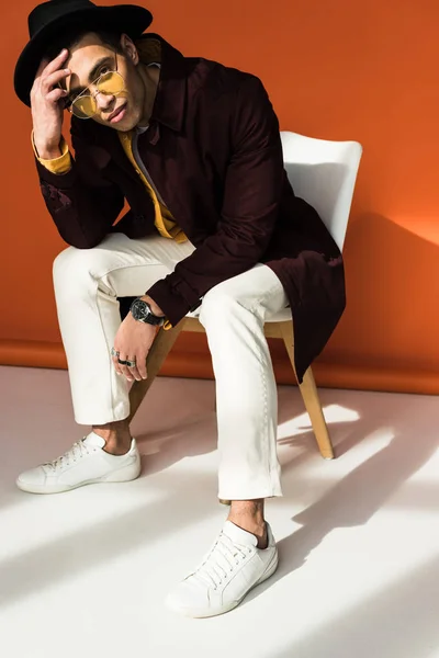 Fashionable mixed race man in hat and sunglasses sitting, looking at camera and posing on orange — Stock Photo