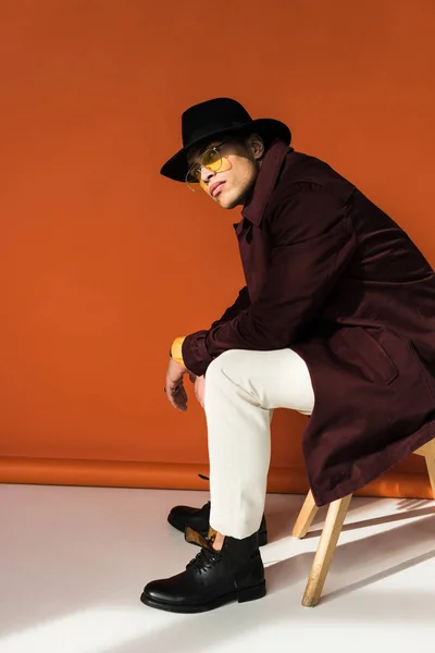 Stylish mixed race man in hat and sunglasses sitting, looking at camera and posing on orange — Stock Photo