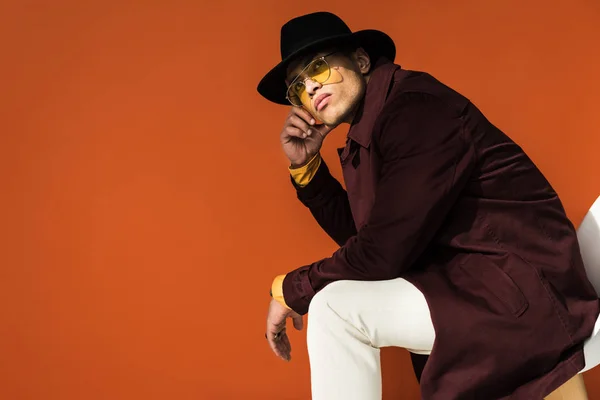 Stylish mixed race man in hat and sunglasses sitting and looking at camera isolated on orange with copy space — Stock Photo