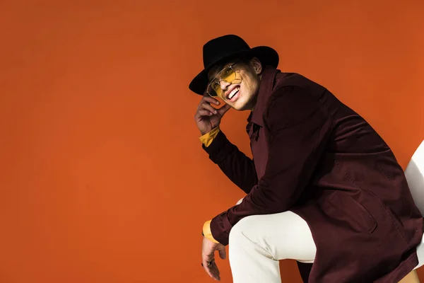 Stylish mixed race man in hat and sunglasses sitting and looking at camera isolated on orange with copy space — Stock Photo