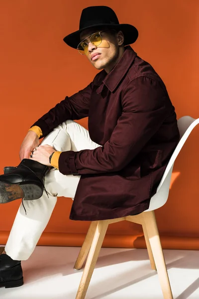 Stylish mixed race man in hat and sunglasses sitting, looking at camera and posing on orange — Stock Photo